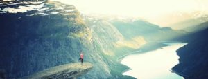 Frau in stehender Yoga-Pose auf Felsvorsprung in großer Höhe in einem Fjord. See und Berge mit Felsen und Wäldern.
